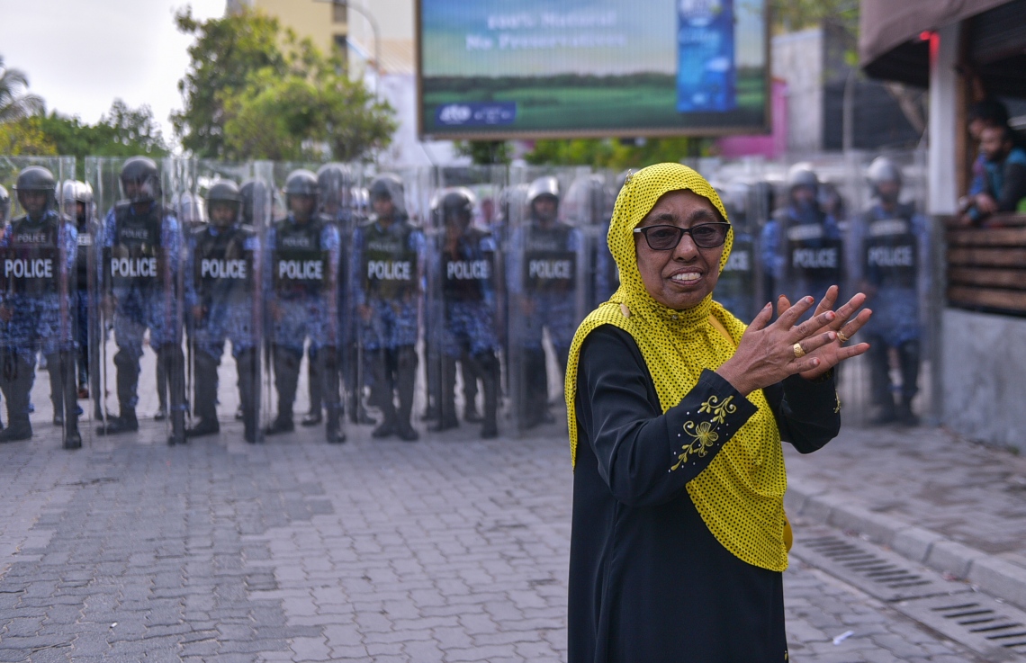 މާލެ: ފެބްރުއަރީ 16، 2018- ސަރުކާރާ ދެކޮޅަށް އިދިކޮޅު ހަރަކާތުން ފަހުގެ ތާރީޚެއްގައި ބާއްވާ ބޮޑު މުޒާހަރާ އަށް ތައްޔާރުވުމަށް "ވޯމްއަޕް މުޒާހަރާ" ގެ ނަމުގައި ކުރިއަށް ގެންދަން އުޅުނު އެއްވުން ރޫޅާލުމަށް އެސްއޯ ފުލުހުން ޝީލްޑާއި މުގުރުބުރިއާއެކު ތައްޔާރަށް ތިބިއިރު، އެމްޑީޕީގެ އަންހެން އެކްޓިވިސްޓަކު މުޒާހަރާގެ ބައިވެރިންނަށް ހިތްވަރު ދީ އަތްޖަހަނީ.---ފޮޓޯ: ހުސެއިން ވަހީދު/މިހާރު