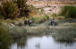 Two competitors carry their bikes as they walk, to cross the river, during stage 6 of the 13th edition of the Titan Desert 2018 mountain biking race between Merzouga and Maadid on May 4, 2018. 
The Titan Desert 2018 is a 600 kilometres mountain bike race completed over six days, snaking between Boumalne Dades, at the foot-slopes of the High Atlas summits, and Erfoud, an oasis town in the Sahara Desert. / AFP PHOTO / FRANCK FIFE