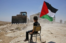 A Palestinian boy sits on a chair with a national flag as Israeli authorities demolish a school site in the village of Yatta, south of the West Bank city of Hebron and to be relocated in another area, on July 11 2018. / AFP PHOTO / HAZEM BADER