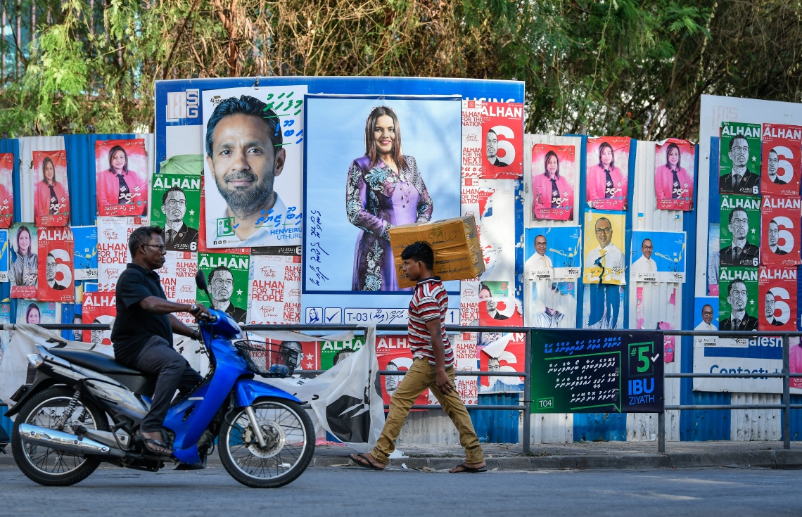މާލޭގެ ކޮންސްޓްރަކްޝަން ސައިޓެއްގެ ބޭރުގައި ގިނަ އަދަދެއްގެ ކެމްޕޭން ޕޯސްޓަރާއި ބިލްބޯޑު ހަރުކޮށްފައި ހުރިއިރު މަގުމަތީގައި މީހުން ހަރަކާތްތެރިވަނީ.---ފޮޓޯ: ނިޝާން އަލީ/މިހާރު