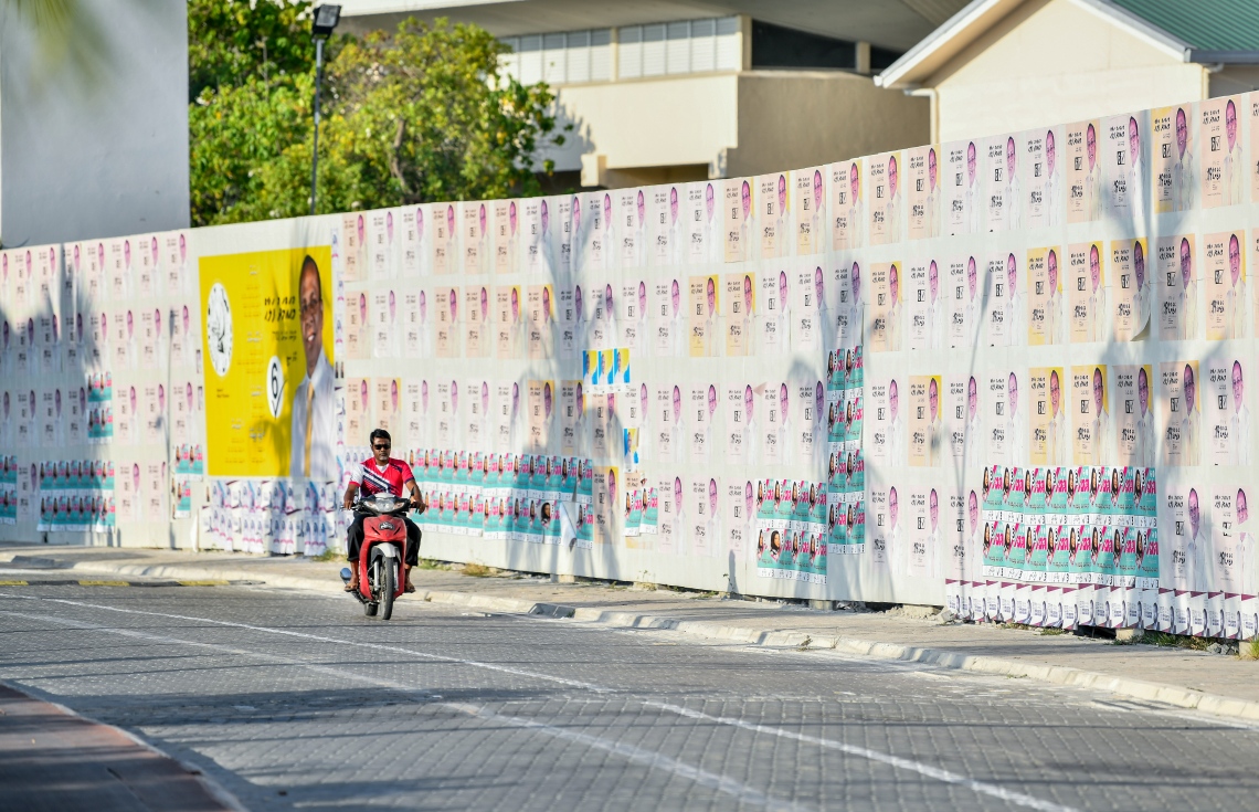 ގިނަ އަދަދެއްގެ ކެމްޕޭން ޕޯސްޓަރު ހަރުކޮށްފައިހުރި ފާރެއްގެ ކައިރިން މީހަކު ސައިކަލެއްގައި ދަނީ.---ފޮޓޯ: ނިޝާން އަލީ/މިހާރު