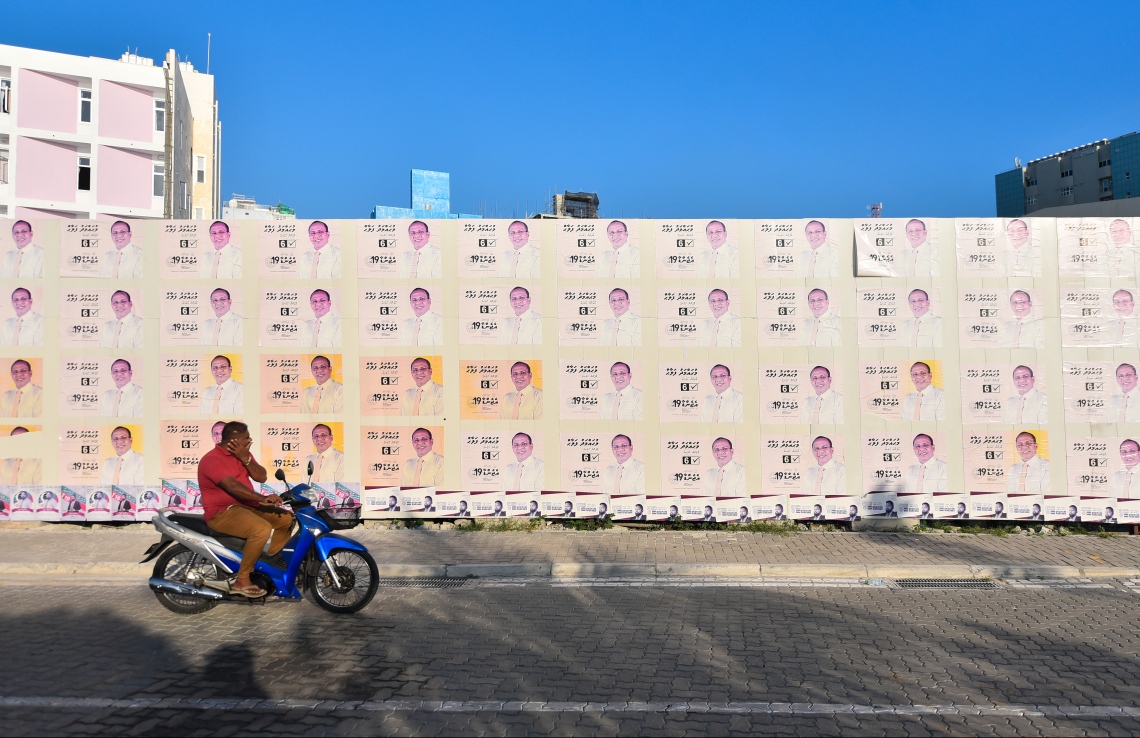 ގިނަ އަދަދެއްގެ ކެމްޕޭން ޕޯސްޓަރު ހަރުކޮށްފައިހުރި ފާރެއްގެ ކައިރިން މީހަކު ސައިކަލެއްގައި ދަނީ.---ފޮޓޯ: ހުސައިން ވަހީދު/މިހާރު