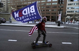 A skateboarder  holds a Biden flag, with a US National falg tied to his neck, ahead of the inauguration ceremonies for President-elect Joe Biden and Vice President-elect Kamala Harris on January 18, 2021 in Washington, DC. - President-elect Joe Biden and Vice President-elect Kamala Harris will be sworn into office January 20, 2021. (Photo by TIMOTHY A. CLARY / AFP)