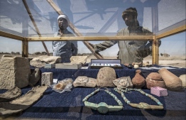 A picture taken on April 10, 2021, shows workers standing next to a display of artifacts uncovered at the archaeological site of a 3000 year old city, dubbed The Rise of Aten, dating to the reign of Amenhotep III, uncovered by the Egyptian mission near Luxor. - Archaeologists have uncovered the remains of an ancient city in the desert outside Luxor that they say is the "largest" ever found in Egypt and dates back to a golden age of the pharaohs 3,000 years ago. Famed Egyptologist Zahi Hawass announced the discovery of the "lost golden city", saying the site was uncovered near Luxor, home of the legendary Valley of the Kings. (Photo by Khaled DESOUKI / AFP)