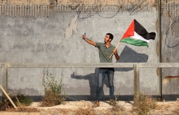 A Palestinian protester lifting a national flag takes a selfie during a demonstration by the border fence with Israel, east of Gaza City, to denounce the Israeli siege of the Palestinian strip and express support for Jerusalem's Al-Aqsa mosque, on August 21, 2021. - Israeli troops fired live rounds at Palestinian protesters who hurled firebombs and burned tyres from behind the Gaza Strip's border fence, with medics reporting 23 Palestinians injured. The protest called for by the Palestinian Hamas movement that rules Gaza marks the burning 52 years ago of Jerusalem's Al-Aqsa Mosque, the third-holiest site in Islam -- Photo: Said Khathib/ AFP