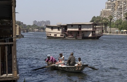 A family of fishermen sits in a boat as one of the houseboats (background) usually moored across one of the banks of the Nile river between the Zamalek district of Egypt's capital Cairo (R) and the Agouza district of its twin city of Giza (L) is towed away by authorities on June 27, 2022, as part of a wider decree to clear all of the river's banks in the area. -- Photo: Khaled Desouki / AFP