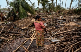 (FILES) A Rohingya woman carries her baby next to her destroyed house at Basara refugee camp in Sittwe on May 16, 2023, after Cyclone Mocha made a landfall. The death toll from Cyclone Mocha that barrelled through Myanmar has reached 145 in the country, the junta's information team said in a statement on May 19. -- Photo: Sai Aung Main / AFP