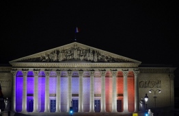 The facade of France's National Assembly is illuminated in French national colours, in Paris, on October 13, 2023, to pay tribute to the teacher killed and two other people severely wounded in a knife attack at a school in the town of Arras in northeastern France. A man of Chechen origin stabbed to death a teacher and severely wounded two other adults on October 13 at a school in northeastern France, with prosectors opening a probe into a suspected act of terror. -- Photo: Geoffroy Van Der Hasselt / AFP